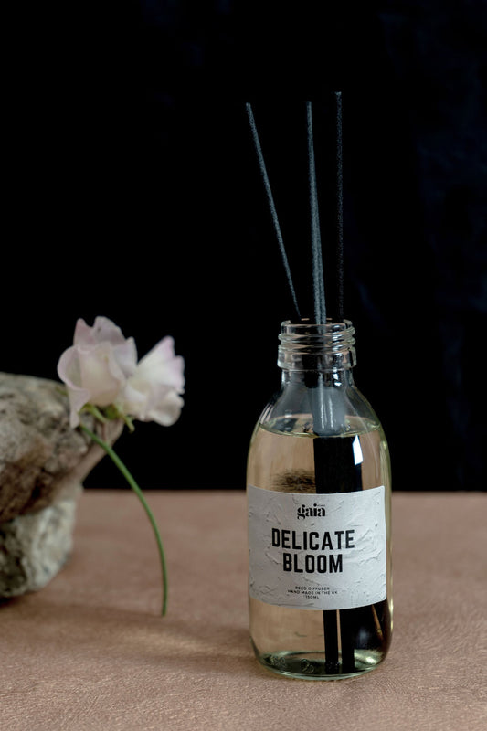 Glass diffuser bottle with four black reeds. Diffuser bottle reads 'Delicate Bloom'